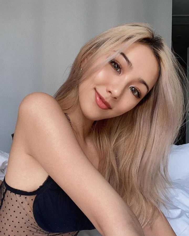 Femme trans aux cheveux blonds, en sous-vêtements noirs, posant sur un lit