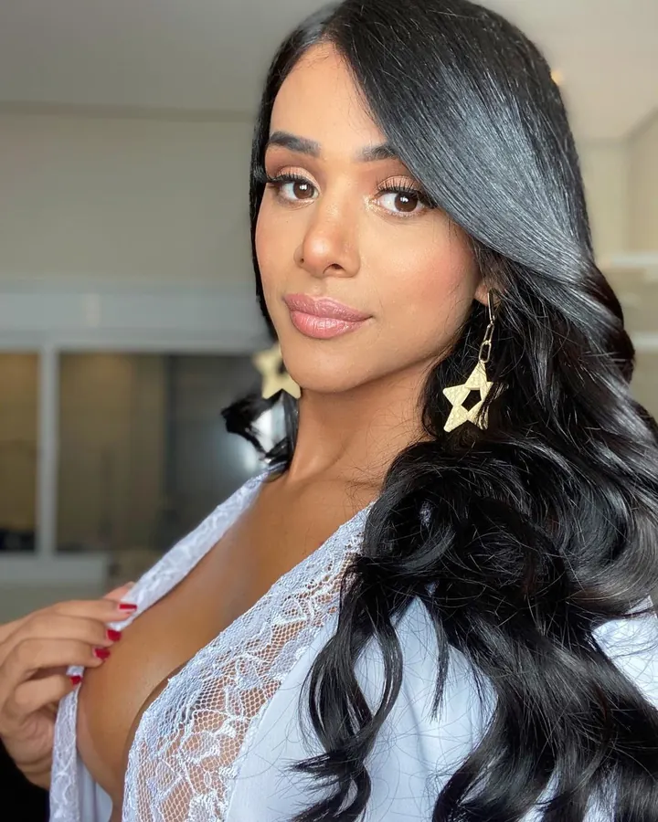 Femme trans latino aux cheveux noirs, seins nus, portant un string floral, tenant un téléphone et regardant la caméra.