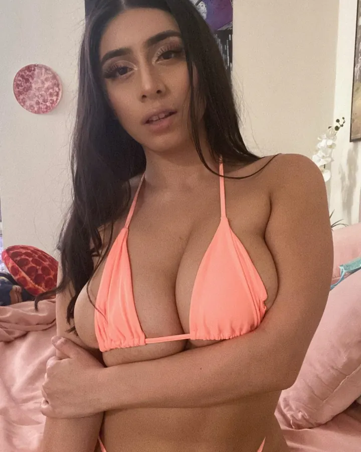 Femme trans européenne aux cheveux bruns, portant un haut noir, se tenant debout dans une chambre, tenant un téléphone pour prendre un selfie.
