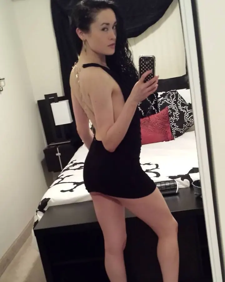 Femme trans en robe noire dos nu, cheveux noirs longs, pose en miroir dans une chambre, regard vers l'objectif.