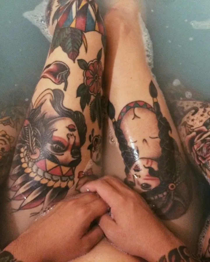 Jambes tatouées d'une femme trans dans un bain moussant, motifs colorés et visages stylisés, mains reposant doucement sur les cuisses.
