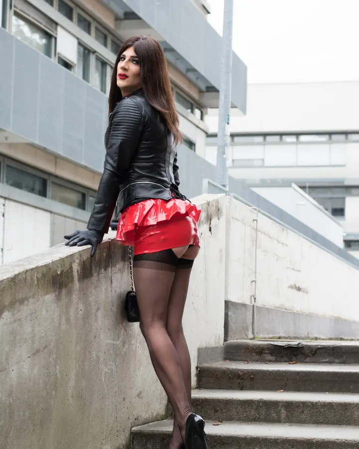 Femme trans aux cheveux bruns, vêtue d'une veste en cuir noir et d'une jupe rouge vernie, posant sur un mur en béton près d'escaliers.