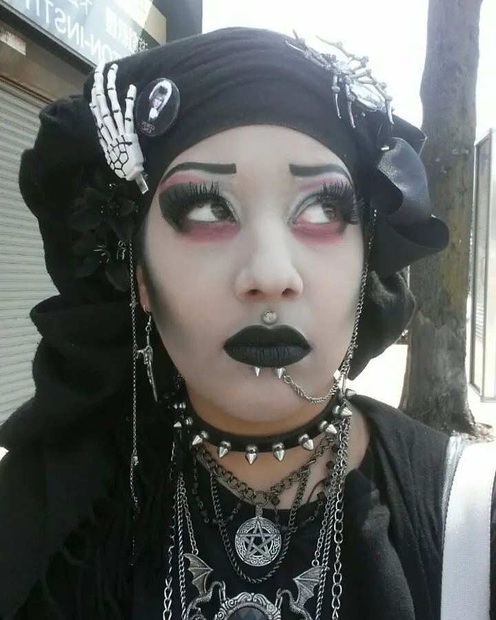 Visage de jeune femme trans avec maquillage gothique, cheveux noirs, peau pâle, portant un bandeau noir orné de bijoux et de chaînes.