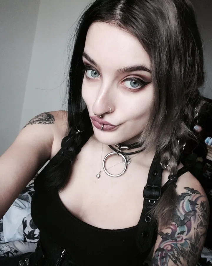 Femme trans aux cheveux bruns, torse nu, posant sur un canapé beige, avec une main sur son entrejambe.