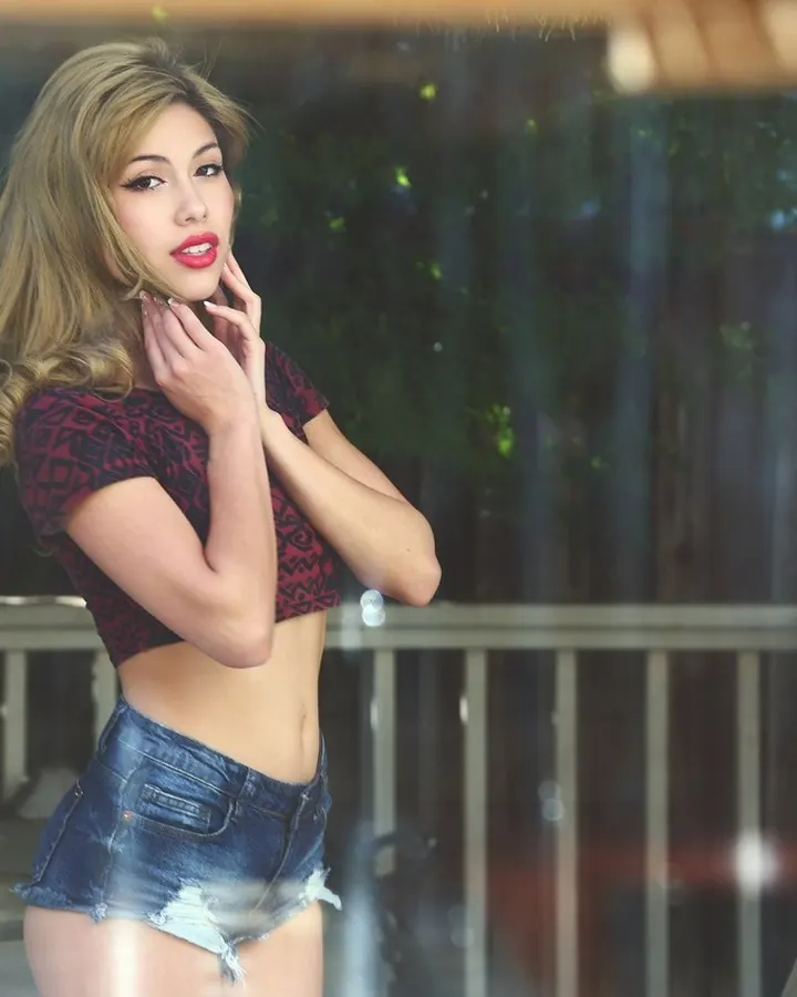 Jeune femme trans aux cheveux blonds, en haut vert clair, posant dans une pièce lumineuse.