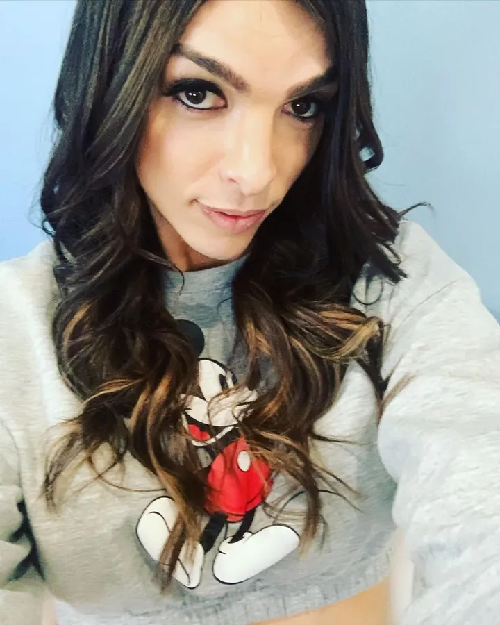 Femme trans aux cheveux bruns portant un sweat Mickey Mouse, regardant directement l'appareil.