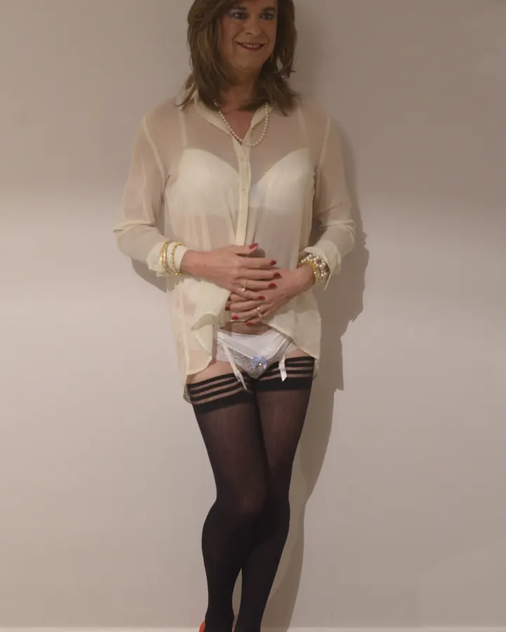 Femme trans aux cheveux châtains, portant une chemise transparente blanche, un soutien-gorge blanc, des collants noirs à rayures et des chaussures rouges, debout contre un mur blanc.