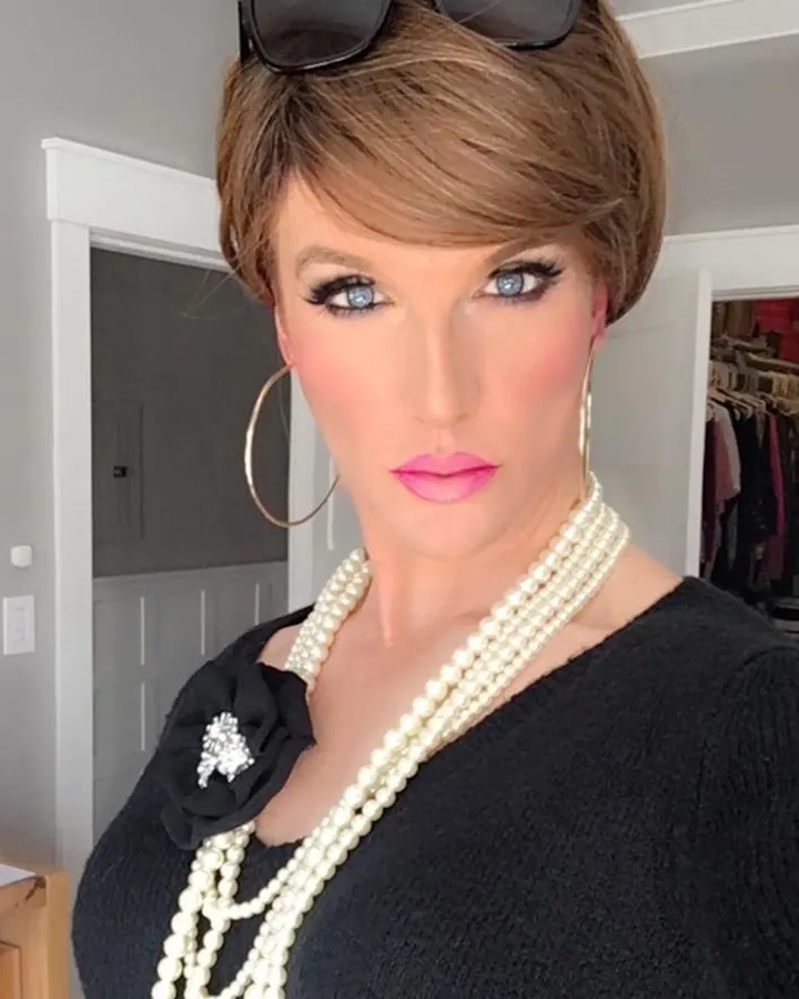 Femme trans aux cheveux bruns courts, portant des lunettes de soleil sur la tête, un pull noir, un collier de perles multiples et une fleur noire sur l'épaule, prenant un selfie.