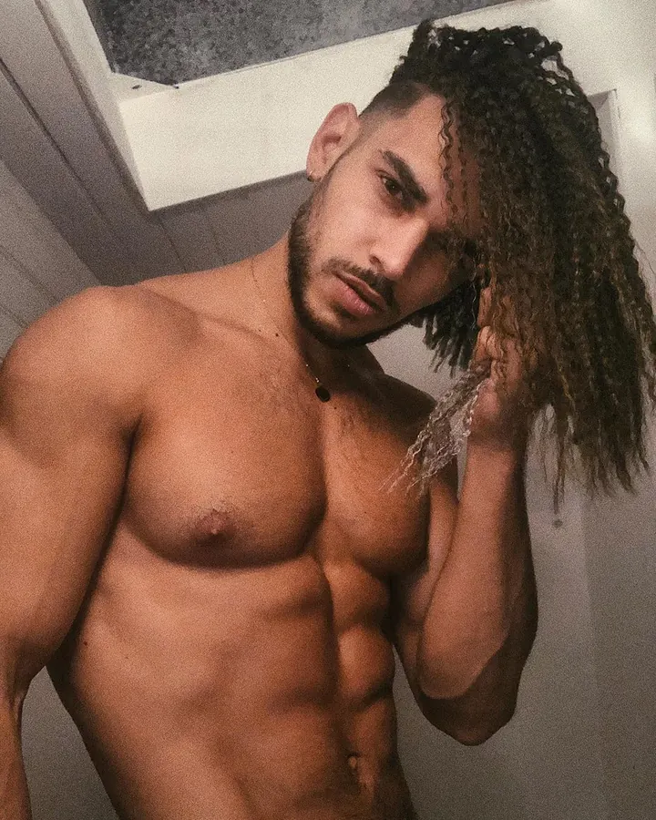 Homme torse nu, aux cheveux bouclés longs sur le côté, avec une barbe courte, tenant une mèche de cheveux, dans un intérieur moderne.