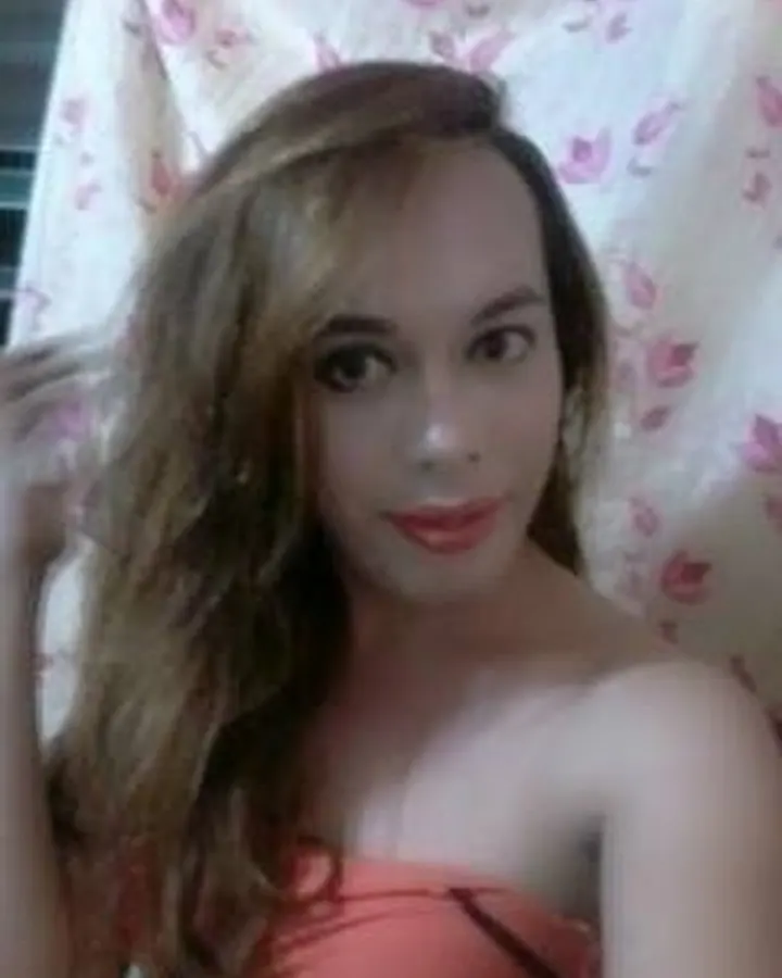 Femme trans aux cheveux bruns, portant un haut orange, devant un rideau à motifs floraux roses et blancs.