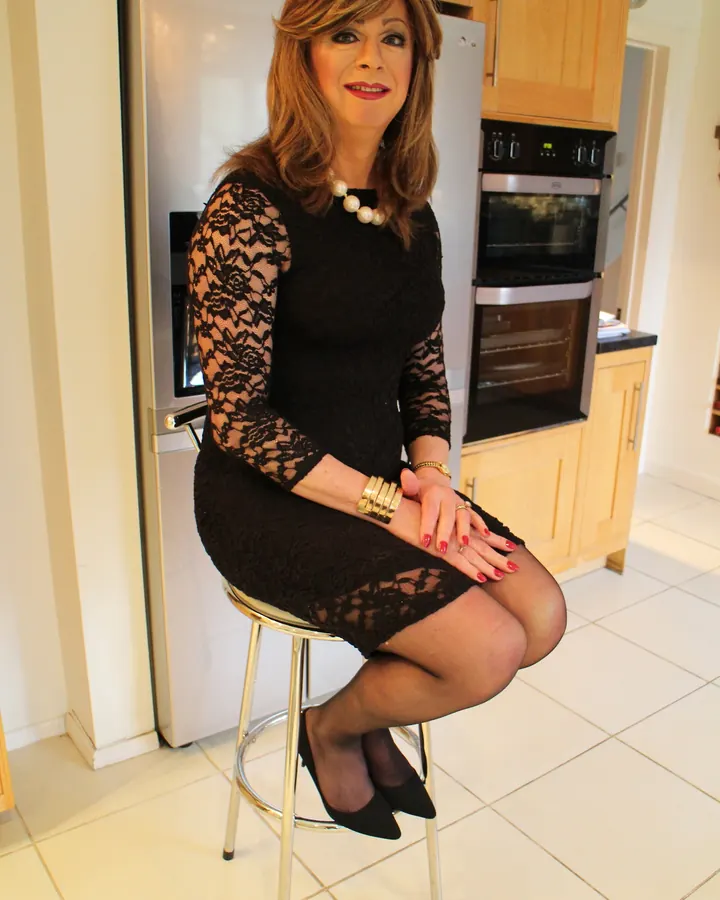 Femme trans aux cheveux blonds, en robe noire dentelle, assise sur un tabouret dans une cuisine moderne.